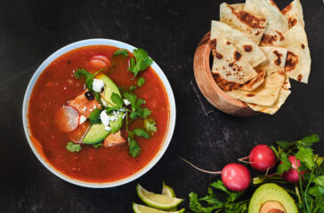 bowl of deep red soup topped with radish, avocado, cheese, and cilantro. surrounded by lime slices and tortilla chips