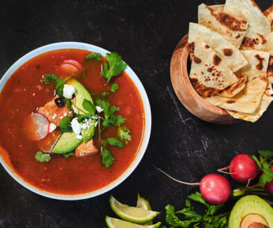 bowl of deep red soup topped with radish, avocado, cheese, and cilantro. surrounded by lime slices and tortilla chips