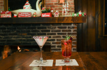 two cocktails on a table in front of the fire. an opaque white drink in a martini glass with red and green sprinkles on the rim. a red drink in a footed highball glass garnished with rosemary