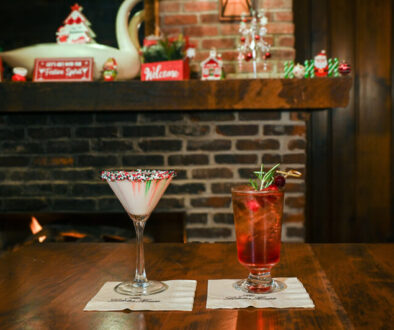 two cocktails on a table in front of the fire. an opaque white drink in a martini glass with red and green sprinkles on the rim. a red drink in a footed highball glass garnished with rosemary